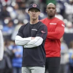 Bobby Slowik, in marineblauem Hemd und Mütze mit verschränkten Armen, steht konzentriert auf dem Fußballfeld. Hinter ihm beobachtet ein weiterer Mann in rotem Hemd die Szene. Der Hintergrund fängt die Energie einer Sportstätte voller verschwommener Zuschauer und Spieler ein. Diese Beschreibung wurde mit der FootballR KI automatisch generiert.