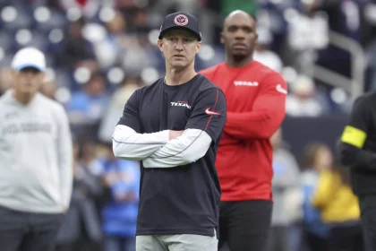Bobby Slowik, in marineblauem Hemd und Mütze mit verschränkten Armen, steht konzentriert auf dem Fußballfeld. Hinter ihm beobachtet ein weiterer Mann in rotem Hemd die Szene. Der Hintergrund fängt die Energie einer Sportstätte voller verschwommener Zuschauer und Spieler ein. Diese Beschreibung wurde mit der FootballR KI automatisch generiert.