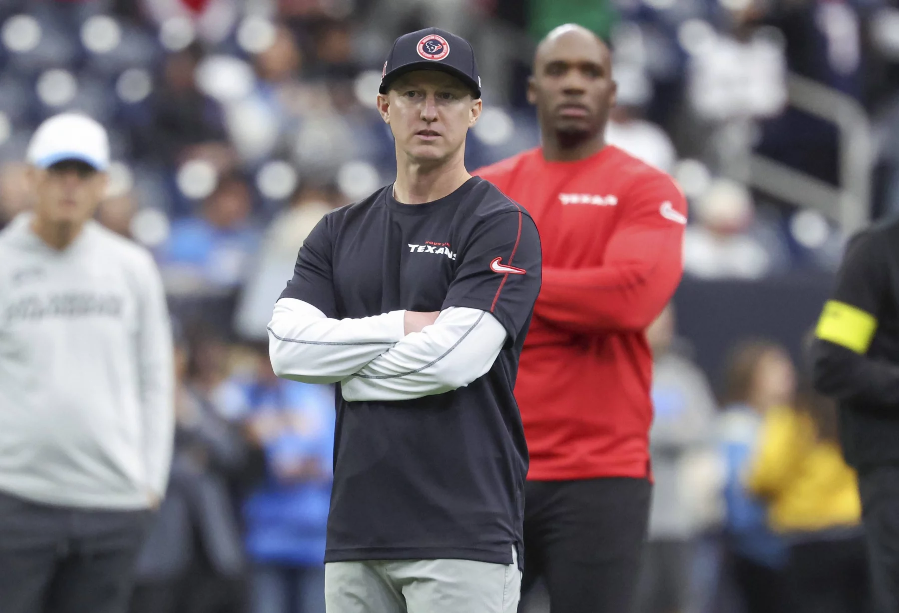 Bobby Slowik, in marineblauem Hemd und Mütze mit verschränkten Armen, steht konzentriert auf dem Fußballfeld. Hinter ihm beobachtet ein weiterer Mann in rotem Hemd die Szene. Der Hintergrund fängt die Energie einer Sportstätte voller verschwommener Zuschauer und Spieler ein. Diese Beschreibung wurde mit der FootballR KI automatisch generiert.
