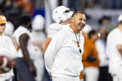 College Football - Steve Sarkisian in einem weißen Kapuzenpullover mit dem Logo der Texas Longhorns steht lächelnd auf einem Footballfeld. Er trägt eine Trillerpfeife um den Hals und die Hände auf dem Rücken. Im Hintergrund sind verschwommen Spieler in weißen Uniformen und Helmen zu sehen. Die Atmosphäre wirkt lebhaft und konzentriert. Diese Beschreibung wurde mit der FootballR KI automatisch generiert.