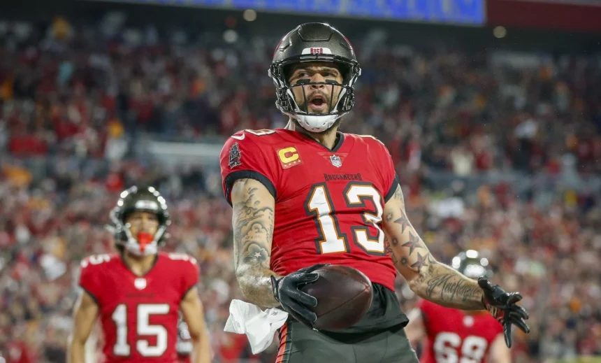 18 Spiele - Footballspieler Mike Evans, der eine rote Uniform der Tampa Bay Buccaneers mit der Nummer 13 trägt, strahlt vor Begeisterung, während er im Stadion einen Football umklammert. Spieler in ähnlichen Uniformen, den Nummern 15 und 69, stehen im Hintergrund, während die verschwommene Menge Evans‘ dynamische Pose hervorhebt. Diese Beschreibung wurde mit der FootballR KI automatisch generiert.