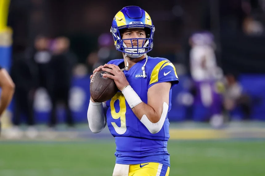 Ein Rams-QB in blau-gelber Uniform, Nummer 9, hält einen Football bereit zum Werfen. Er trägt einen blauen Helm mit gelben Akzenten und steht auf dem Spielfeld. Die verschwommenen Figuren und Stadionlichter im Hintergrund spiegeln die Intensität von Matthew Staffords Konzentration am Spieltag wider. Diese Beschreibung wurde mit der FootballR KI automatisch generiert.