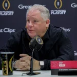 Chip Kelly mit kurzen grauen Haaren, der als „Top-Verdiener“ Chip Kelly identifiziert wird, sitzt an einem Tisch mit einem Mikrofon mit der Aufschrift „CFB“. Er trägt eine schwarze Jacke vor einem Hintergrund mit den Logos „College Football Playoff“ und „Great Clips“. Neben ihm liegen ein bunter Reisebecher und ein Stift. Diese Beschreibung wurde mit der FootballR KI automatisch generiert.