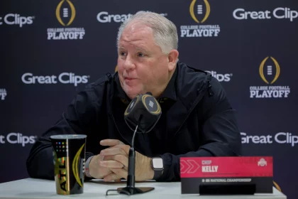 Chip Kelly mit kurzen grauen Haaren, der als „Top-Verdiener“ Chip Kelly identifiziert wird, sitzt an einem Tisch mit einem Mikrofon mit der Aufschrift „CFB“. Er trägt eine schwarze Jacke vor einem Hintergrund mit den Logos „College Football Playoff“ und „Great Clips“. Neben ihm liegen ein bunter Reisebecher und ein Stift. Diese Beschreibung wurde mit der FootballR KI automatisch generiert.