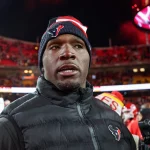 DeMeco Ryans mit einer Mütze der Houston Texans und einer schwarzen Jacke steht auf dem Footballfeld. Das Stadion ist voller Zuschauer auf roten Sitzen, ihre Aufregung ist spürbar. Unter DeMeco Ryans wachsamem Auge ist im Hintergrund eine große Anzeigetafel vor einem weitgehend klaren Nachthimmel zu sehen. Diese Beschreibung wurde mit der FootballR KI automatisch generiert.