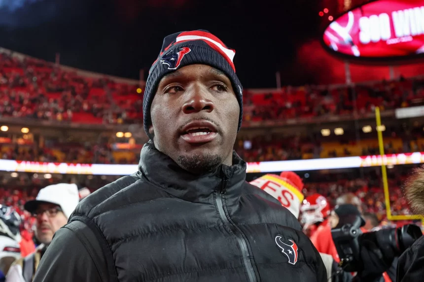 DeMeco Ryans mit einer Mütze der Houston Texans und einer schwarzen Jacke steht auf dem Footballfeld. Das Stadion ist voller Zuschauer auf roten Sitzen, ihre Aufregung ist spürbar. Unter DeMeco Ryans wachsamem Auge ist im Hintergrund eine große Anzeigetafel vor einem weitgehend klaren Nachthimmel zu sehen. Diese Beschreibung wurde mit der FootballR KI automatisch generiert.