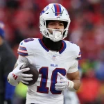 Der Footballspieler der Buffalo Bills, Khalil Shakir, trägt eine weiße Uniform mit der Nummer 10. Er hält einen Football selbstbewusst in der Hand, trägt einen schwarzen Halsschutz und weiße Handschuhe. Das ikonische Bills-Logo leuchtet auf seinem Helm, während im Hintergrund die verschwommene Menge im Stadion summt. Diese Beschreibung wurde mit der FootballR KI automatisch generiert.