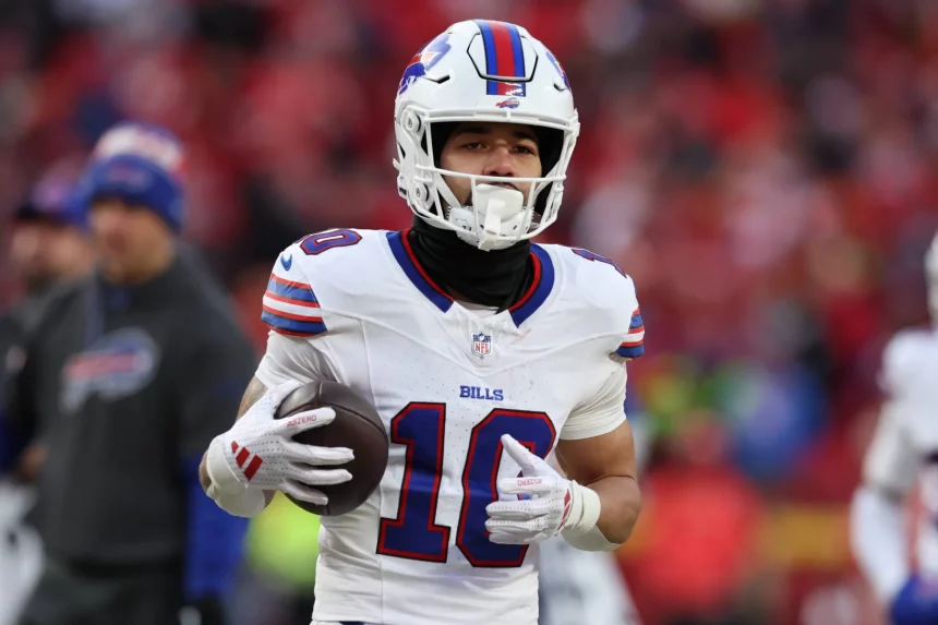 Der Footballspieler der Buffalo Bills, Khalil Shakir, trägt eine weiße Uniform mit der Nummer 10. Er hält einen Football selbstbewusst in der Hand, trägt einen schwarzen Halsschutz und weiße Handschuhe. Das ikonische Bills-Logo leuchtet auf seinem Helm, während im Hintergrund die verschwommene Menge im Stadion summt. Diese Beschreibung wurde mit der FootballR KI automatisch generiert.