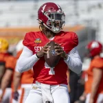 Jalen Milroe, in einem roten Trikot mit der Nummer 4, übt das Werfen eines Passes. Er trägt einen roten Helm mit einer weißen Gesichtsmaske und hält einen braunen Football in der Hand. Er fällt auf, während andere Spieler in orangefarbenen Trikots und ein leeres Stadion im Hintergrund stehen. Diese Beschreibung wurde mit der FootballR KI automatisch generiert.