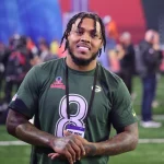 Josh Jacobs in einem dunkelgrünen Sporttrikot mit der Nummer 8, der an einen Top-Receiver erinnert, lächelt und steht auf einem Footballfeld. Er hat geflochtene Haare und Tattoos auf seinem linken Arm. Um seinen Hals hängt ein schwarzes Schlüsselband mit der Aufschrift „PRO BOWL“. Im Hintergrund sind verschwommene Menschen und bunte Banner zu sehen, die den Geist von Green Bay heraufbeschwören. Diese Beschreibung wurde mit der FootballR KI automatisch generiert.