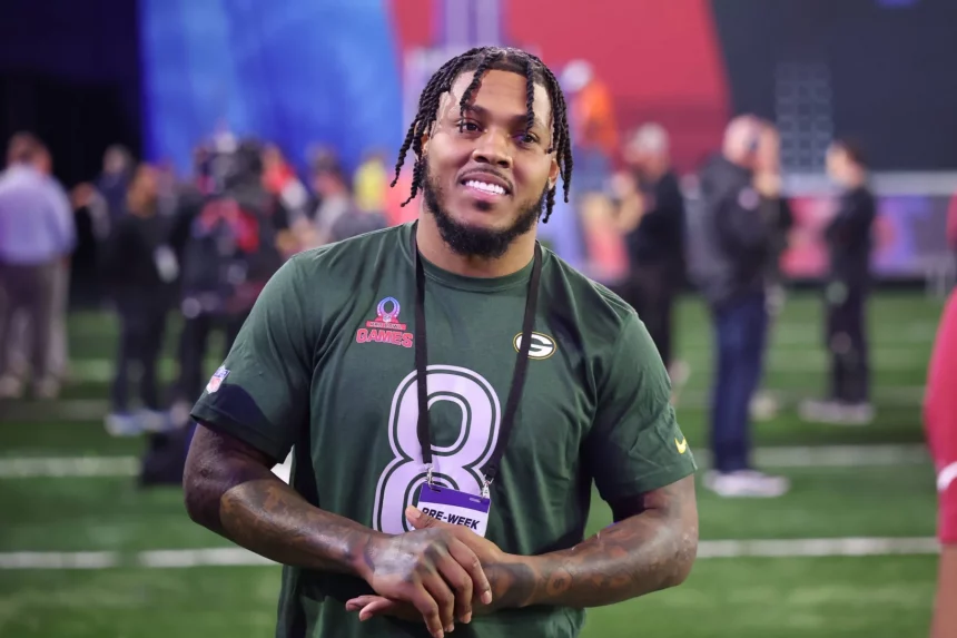 Josh Jacobs in einem dunkelgrünen Sporttrikot mit der Nummer 8, der an einen Top-Receiver erinnert, lächelt und steht auf einem Footballfeld. Er hat geflochtene Haare und Tattoos auf seinem linken Arm. Um seinen Hals hängt ein schwarzes Schlüsselband mit der Aufschrift „PRO BOWL“. Im Hintergrund sind verschwommene Menschen und bunte Banner zu sehen, die den Geist von Green Bay heraufbeschwören. Diese Beschreibung wurde mit der FootballR KI automatisch generiert.