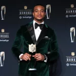 Patrick Surtain in einem grünen Samtanzug und einer schwarzen Fliege, Patrick Surtain, hält eine NFL-Trophäe auf einer Bühne mit schwarzem Hintergrund, auf dem die NFL Honors- und Invisalign-Logos zu sehen sind. Er steht selbstbewusst in der Bildmitte und hat ordentlich gestylte Haare. Diese Beschreibung wurde mit der FootballR KI automatisch generiert.
