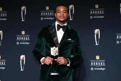 Patrick Surtain in einem grünen Samtanzug und einer schwarzen Fliege, Patrick Surtain, hält eine NFL-Trophäe auf einer Bühne mit schwarzem Hintergrund, auf dem die NFL Honors- und Invisalign-Logos zu sehen sind. Er steht selbstbewusst in der Bildmitte und hat ordentlich gestylte Haare. Diese Beschreibung wurde mit der FootballR KI automatisch generiert.