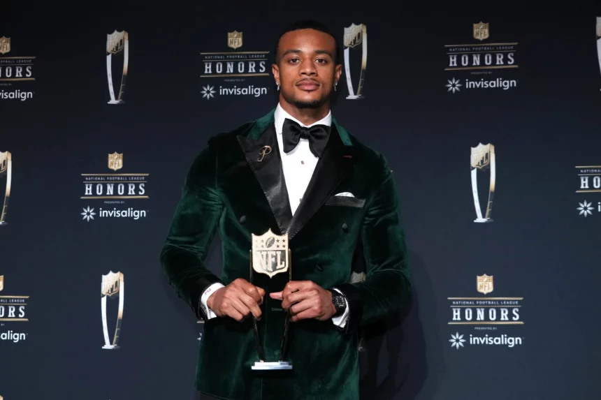 Patrick Surtain in einem grünen Samtanzug und einer schwarzen Fliege, Patrick Surtain, hält eine NFL-Trophäe auf einer Bühne mit schwarzem Hintergrund, auf dem die NFL Honors- und Invisalign-Logos zu sehen sind. Er steht selbstbewusst in der Bildmitte und hat ordentlich gestylte Haare. Diese Beschreibung wurde mit der FootballR KI automatisch generiert.