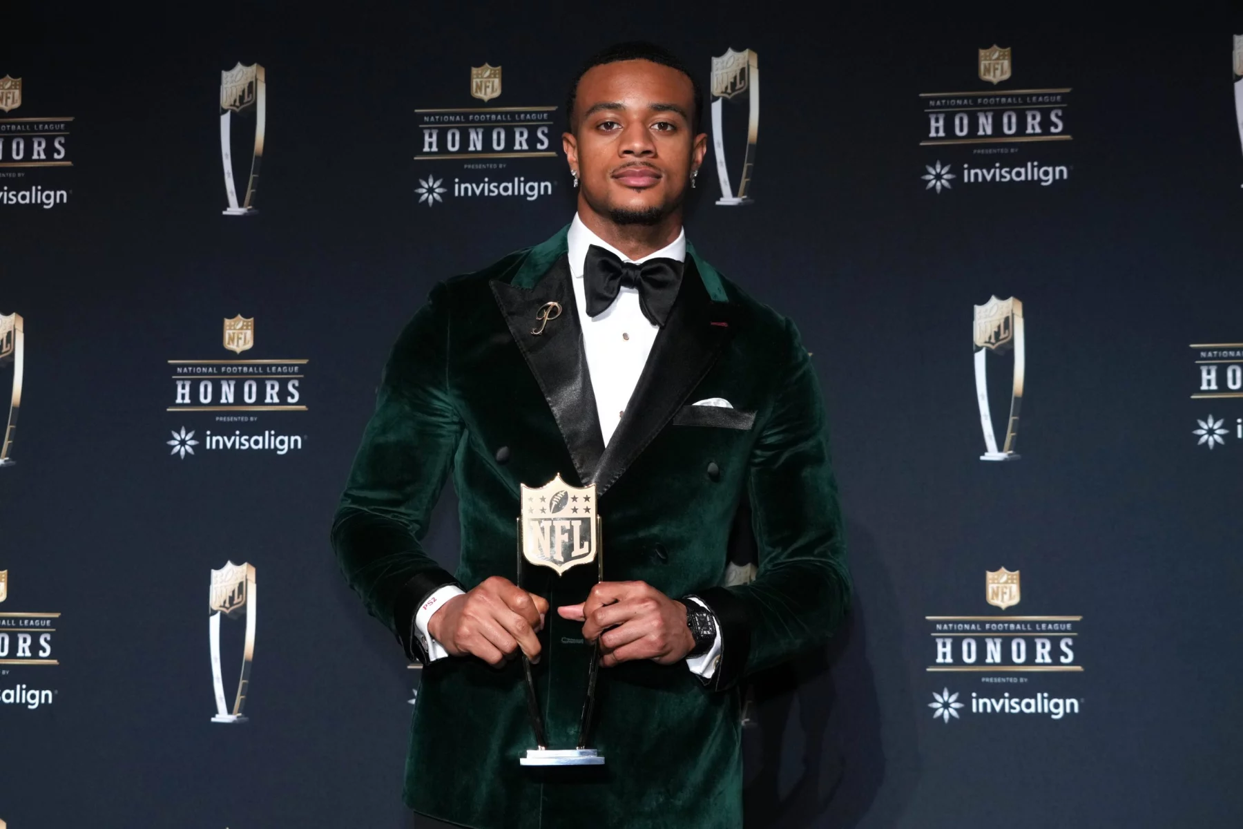 Patrick Surtain in einem grünen Samtanzug und einer schwarzen Fliege, Patrick Surtain, hält eine NFL-Trophäe auf einer Bühne mit schwarzem Hintergrund, auf dem die NFL Honors- und Invisalign-Logos zu sehen sind. Er steht selbstbewusst in der Bildmitte und hat ordentlich gestylte Haare. Diese Beschreibung wurde mit der FootballR KI automatisch generiert.