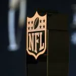 NFL Schedule 2025 - Salary Cap 2025 - Nahaufnahme eines bronzefarbenen NFL-Schildlogos auf einem schwarzen Trophäensockel. Das Emblem mit einem Football und acht Sternen ragt im Vordergrund hervor. Im dunklen, verschwommenen Hintergrund weisen Andeutungen von weißem Text in die Zukunft – möglicherweise als Hinweis auf die Gehaltsobergrenze 2025 und ihre bevorstehenden Auswirkungen. Diese Beschreibung wurde mit der FootballR KI automatisch generiert.