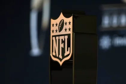 NFL Schedule 2025 - Salary Cap 2025 - Cap Space - Nahaufnahme eines bronzefarbenen NFL-Schildlogos auf einem schwarzen Trophäensockel. Das Emblem mit einem Football und acht Sternen ragt im Vordergrund hervor. Im dunklen, verschwommenen Hintergrund weisen Andeutungen von weißem Text in die Zukunft – möglicherweise als Hinweis auf die Gehaltsobergrenze 2025 und ihre bevorstehenden Auswirkungen. Diese Beschreibung wurde mit der FootballR KI automatisch generiert.