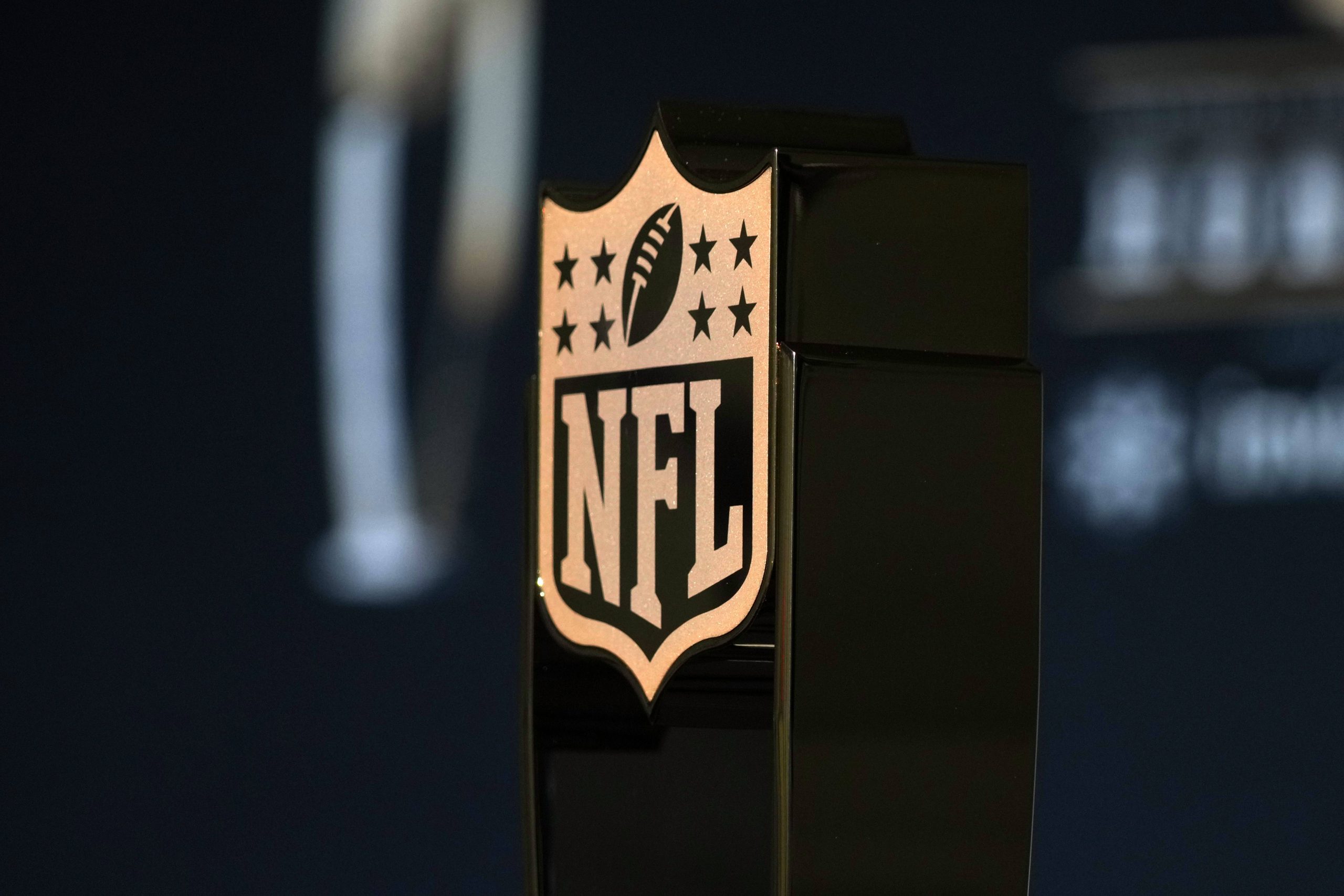 NFL Schedule 2025 - Salary Cap 2025 - Nahaufnahme eines bronzefarbenen NFL-Schildlogos auf einem schwarzen Trophäensockel. Das Emblem mit einem Football und acht Sternen ragt im Vordergrund hervor. Im dunklen, verschwommenen Hintergrund weisen Andeutungen von weißem Text in die Zukunft – möglicherweise als Hinweis auf die Gehaltsobergrenze 2025 und ihre bevorstehenden Auswirkungen. Diese Beschreibung wurde mit der FootballR KI automatisch generiert.