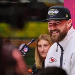 Travis Kelce in einer weißen Reißverschlussjacke mit einem Logo der Kansas City Chiefs lächelt, während er während eines Chiefs Update in ein Mikrofon des NFL Network spricht. Er trägt eine schwarze Kappe mit einem „Super Bowl LVII“-Aufnäher. Ein buntes abstraktes Design verwischt den Hintergrund, in dem neben ihm eine Frau teilweise sichtbar ist. Diese Beschreibung wurde mit der FootballR KI automatisch generiert.