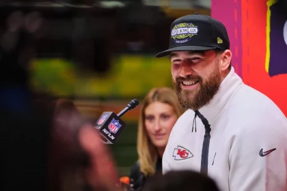 Travis Kelce in einer weißen Reißverschlussjacke mit einem Logo der Kansas City Chiefs lächelt, während er während eines Chiefs Update in ein Mikrofon des NFL Network spricht. Er trägt eine schwarze Kappe mit einem „Super Bowl LVII“-Aufnäher. Ein buntes abstraktes Design verwischt den Hintergrund, in dem neben ihm eine Frau teilweise sichtbar ist. Diese Beschreibung wurde mit der FootballR KI automatisch generiert.