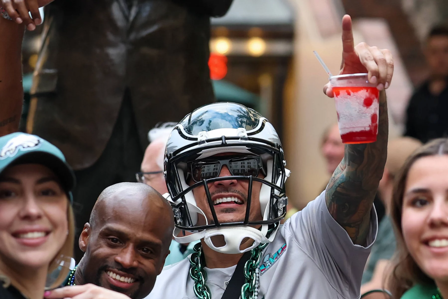 NFC East Draft Bewertung - Eine Person mit einem Philadelphia Eagles Helm lächelt und zeigt nach oben. Sie hält ein Getränk mit roten und weißen Schichten in der Hand. Umgeben von anderen Eagles-Fans erinnert die festliche Szene an die Feierlichkeiten beim Super Bowl LIX in New Orleans, bei dem jeder für das große Spiel in der Ausrüstung seines Teams gekleidet ist. Diese Beschreibung wurde mit der FootballR KI automatisch generiert.