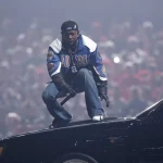 Kendrick Lamar hockt auf einem Autodach. Er trägt eine blau-weiße Jacke, schwarze Hosen und eine verkehrt herum aufgesetzte Kappe. Die Fans jubeln wild in der dynamischen Konzertszene, die an die Halbzeitshow des Super Bowl erinnert. Scheinwerfer- und Raucheffekte verleihen diesem Moment, der ein Rekord sein könnte, noch mehr Dramatik. Diese Beschreibung wurde mit der FootballR KI automatisch generiert.