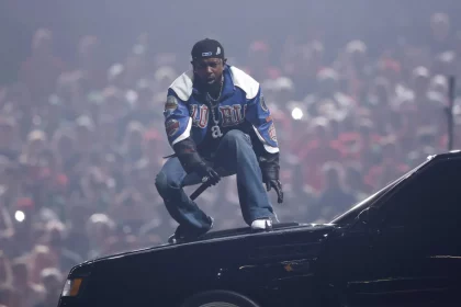 Kendrick Lamar hockt auf einem Autodach. Er trägt eine blau-weiße Jacke, schwarze Hosen und eine verkehrt herum aufgesetzte Kappe. Die Fans jubeln wild in der dynamischen Konzertszene, die an die Halbzeitshow des Super Bowl erinnert. Scheinwerfer- und Raucheffekte verleihen diesem Moment, der ein Rekord sein könnte, noch mehr Dramatik. Diese Beschreibung wurde mit der FootballR KI automatisch generiert.