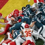 Tush Push - Spieler zweier American-Football-Teams, eines in dunkelgrünen Uniformen, die an die Green Bay Packers erinnern, und das andere in weiß und rot, drängen und schubsen in einem dicht gedrängten Gedränge in der Nähe der Endzone. Der Fokus liegt auf einem „Tush Push“-Spiel mit intensivem Körperkontakt, während sie in der Nähe der Torlinie um den Ballbesitz kämpfen. Diese Beschreibung wurde mit der FootballR KI automatisch generiert.