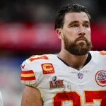 Travis Kelce mit Bart steht auf dem Feld und trägt ein weißes Trikot mit roten und gelben Akzenten. Das Trikot zeigt die Nummer 87, Teamlogos, ein Kapitänsabzeichen und ein Super-Bowl-Abzeichen. Er wirkt konzentriert, im Hintergrund ist eine verschwommene Menschenmenge zu sehen. Diese Beschreibung wurde mit der FootballR KI automatisch generiert.