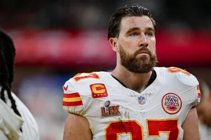 Travis Kelce mit Bart steht auf dem Feld und trägt ein weißes Trikot mit roten und gelben Akzenten. Das Trikot zeigt die Nummer 87, Teamlogos, ein Kapitänsabzeichen und ein Super-Bowl-Abzeichen. Er wirkt konzentriert, im Hintergrund ist eine verschwommene Menschenmenge zu sehen. Diese Beschreibung wurde mit der FootballR KI automatisch generiert.
