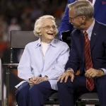 Virginia McCaskey mit kurzen weißen Haaren, identifiziert als Virginia McCaskey von den Chicago Bears, sitzt lächelnd neben einem Mann in einem blauen Anzug mit roter Krawatte. Sie sitzen auf der Bühne mit einem verschwommenen Publikum im Hintergrund und scheinen ein freundliches Gespräch zu führen. Diese Beschreibung wurde mit der FootballR KI automatisch generiert.