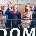 Eine Gruppe von Menschen steht in einer Stadionloge. US Präsident Donald Trump im dunklen Anzug salutiert, während andere ihre Hände aufs Herz legen. Eine Frau in Weiß steht daneben. Vorne ahmt ein Kind die Geste nach. Im Hintergrund ist ein Teil eines Schildes mit der Aufschrift „DOME“ zu sehen. Diese Beschreibung wurde mit der FootballR KI automatisch generiert.