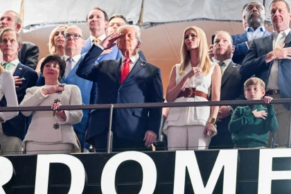 Eine Gruppe von Menschen steht in einer Stadionloge. US Präsident Donald Trump im dunklen Anzug salutiert, während andere ihre Hände aufs Herz legen. Eine Frau in Weiß steht daneben. Vorne ahmt ein Kind die Geste nach. Im Hintergrund ist ein Teil eines Schildes mit der Aufschrift „DOME“ zu sehen. Diese Beschreibung wurde mit der FootballR KI automatisch generiert.