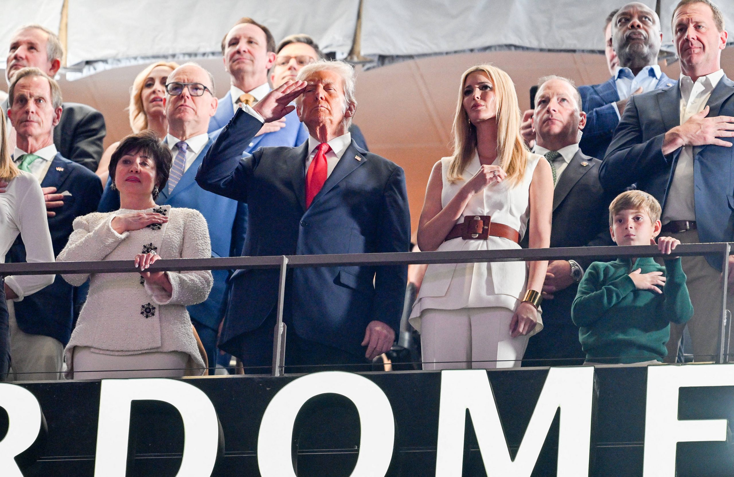 Eine Gruppe von Menschen steht in einer Stadionloge. US Präsident Donald Trump im dunklen Anzug salutiert, während andere ihre Hände aufs Herz legen. Eine Frau in Weiß steht daneben. Vorne ahmt ein Kind die Geste nach. Im Hintergrund ist ein Teil eines Schildes mit der Aufschrift „DOME“ zu sehen. Diese Beschreibung wurde mit der FootballR KI automatisch generiert.