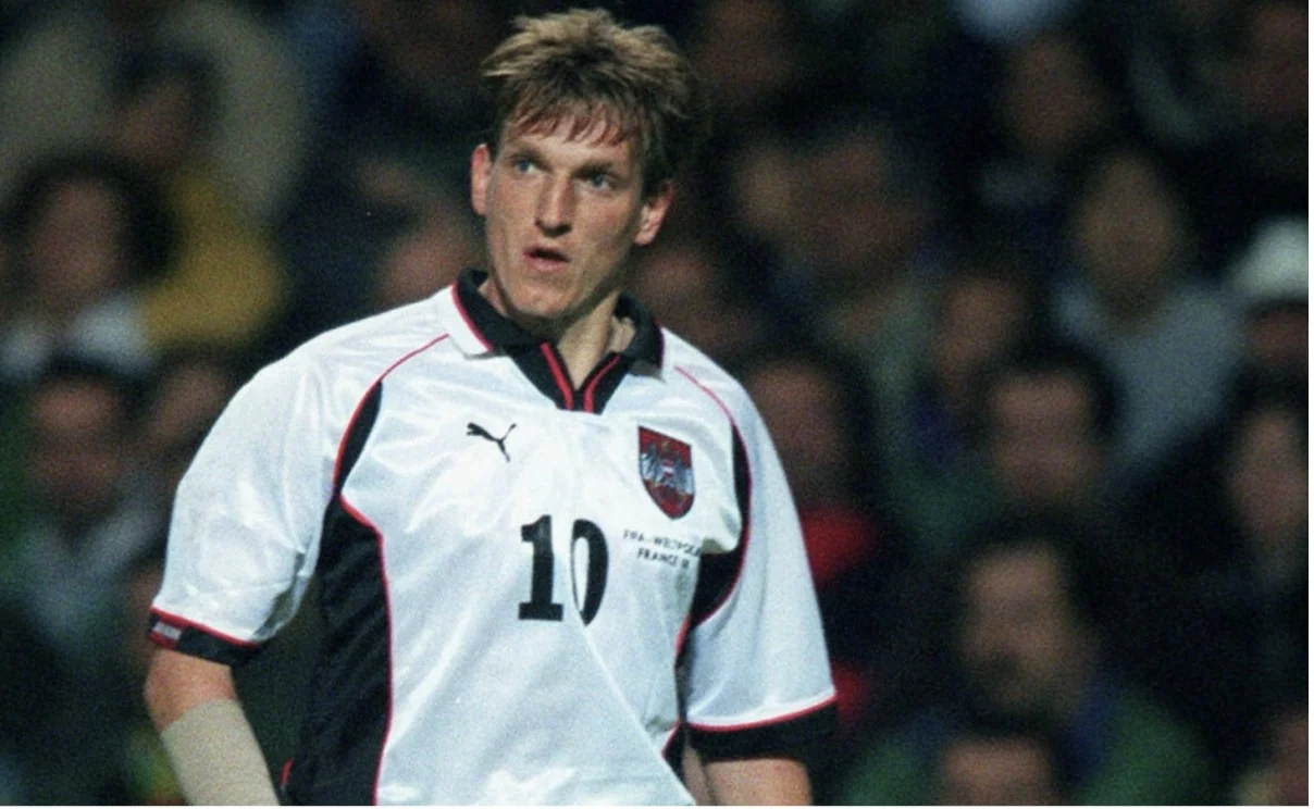 Ein Fußballspieler in einem weißen Trikot mit schwarzen und roten Akzenten steht auf dem Feld. Das Trikot trägt ein Puma-Logo, mit „10“ auf der Vorderseite und ein Emblem. Der Spieler ist einer der erfolgreichsten österreichischen Fußballer und wirkt konzentriert, während Zuschauer in dunkler Kleidung im Hintergrund verschwimmen. Diese Beschreibung wurde mit der FootballR KI automatisch generiert.