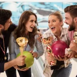 Vier Personen feiern fröhlich auf einer Bowlingbahn und halten jeweils einen Pokal in den Händen. Zwei Männer und zwei Frauen stehen dicht beieinander, lächeln und lachen. Mit einer Mischung aus Spaß und Strategie hält eine Frau eine grüne Bowlingkugel, die andere eine rosa. Vor dem Hintergrund der verschwommenen Bowlingbahnen und -kegel. Diese Beschreibung wurde mit der FootballR KI automatisch generiert.