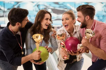 Vier Personen feiern fröhlich auf einer Bowlingbahn und halten jeweils einen Pokal in den Händen. Zwei Männer und zwei Frauen stehen dicht beieinander, lächeln und lachen. Mit einer Mischung aus Spaß und Strategie hält eine Frau eine grüne Bowlingkugel, die andere eine rosa. Vor dem Hintergrund der verschwommenen Bowlingbahnen und -kegel. Diese Beschreibung wurde mit der FootballR KI automatisch generiert.