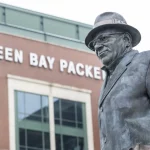 432,6 Millionen Dollar - Die Statue eines Mannes in Anzug und Fedora steht vor einem Gebäude mit der Aufschrift „GREEN BAY PACKERS“ in fetten weißen Buchstaben. Die Backsteinfassade und die großen Fenster lassen auf den Eingang eines Stadions oder einer Anlage schließen. Die Statue scheint an eine bedeutende Persönlichkeit der Sportgeschichte zu erinnern. Diese Beschreibung wurde mit der FootballR KI automatisch generiert.