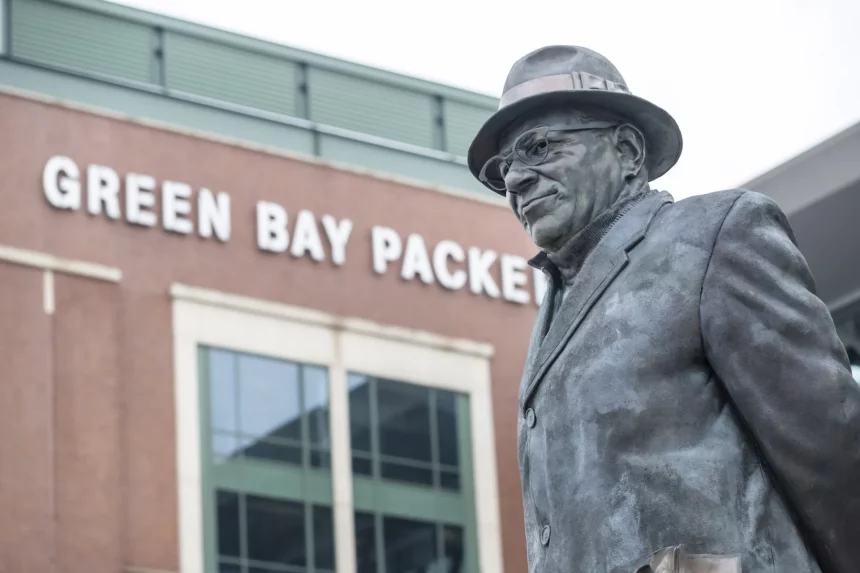 432,6 Millionen Dollar - Die Statue eines Mannes in Anzug und Fedora steht vor einem Gebäude mit der Aufschrift „GREEN BAY PACKERS“ in fetten weißen Buchstaben. Die Backsteinfassade und die großen Fenster lassen auf den Eingang eines Stadions oder einer Anlage schließen. Die Statue scheint an eine bedeutende Persönlichkeit der Sportgeschichte zu erinnern. Diese Beschreibung wurde mit der FootballR KI automatisch generiert.