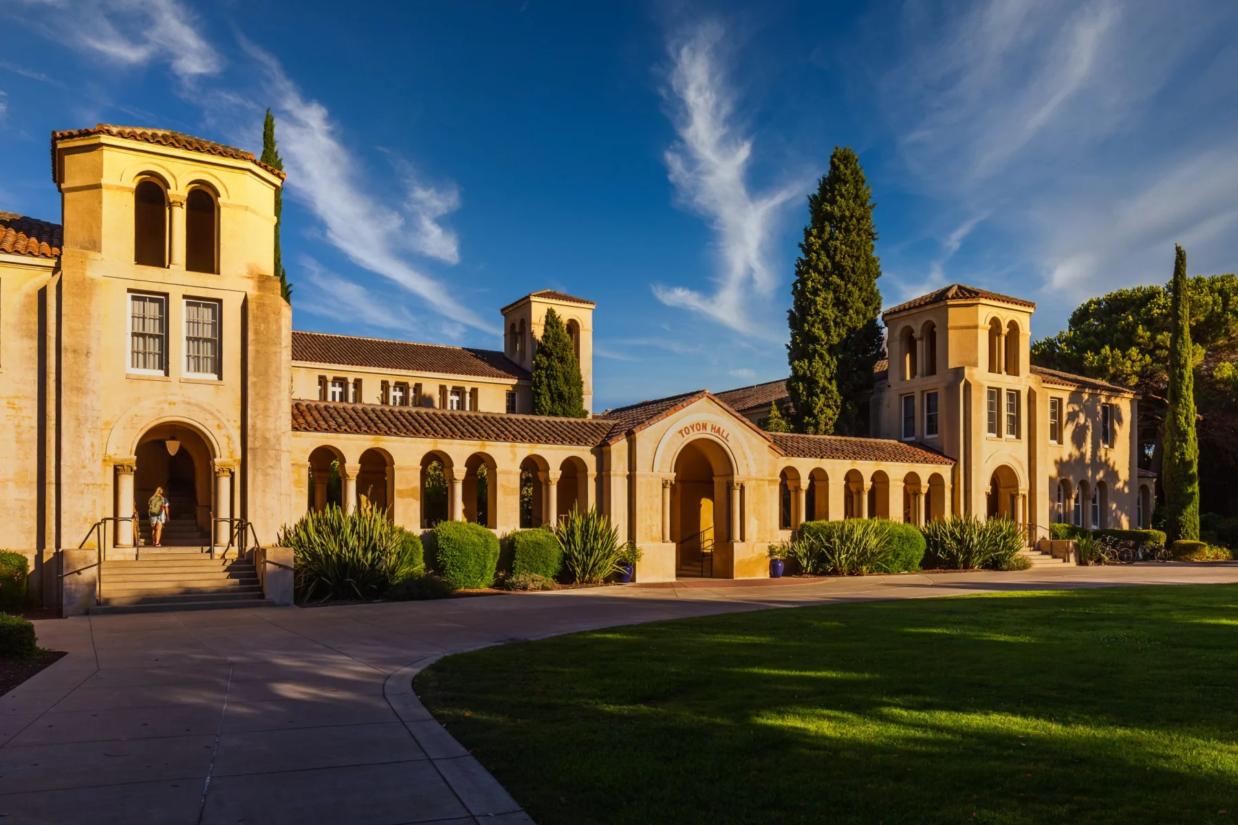 Stanford University enthlässt HC Troy Taylor - Ein Gebäude im spanischen Stil mit gewölbten Gängen und Ziegeldach sonnt sich im warmen Licht des Sonnenuntergangs. Die Fassade erinnert an Stanfords ikonische Architektur und besticht durch Säulen und kunstvolle Schnitzereien. Eine Person steigt die Stufen hinauf, während üppiges Grün diese ruhige Szene unter einem klaren blauen Himmel umgibt. Diese Beschreibung wurde mit der FootballR KI automatisch generiert.