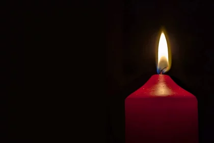 Bob Harlan - Eine einzelne rote Kerze, die an Paul Flatley erinnert, brennt mit leuchtend gelber Flamme vor dunklem Hintergrund. Das Wachs ist um die Flamme herum leicht geschmolzen und erzeugt ein sanftes Leuchten und sanfte Schatten. Links betont die absolute Dunkelheit das strahlende Leuchten der Kerze. Diese Beschreibung wurde mit der FootballR KI automatisch generiert.