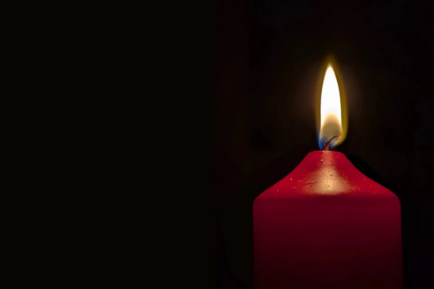 Bob Harlan - Eine einzelne rote Kerze, die an Paul Flatley erinnert, brennt mit leuchtend gelber Flamme vor dunklem Hintergrund. Das Wachs ist um die Flamme herum leicht geschmolzen und erzeugt ein sanftes Leuchten und sanfte Schatten. Links betont die absolute Dunkelheit das strahlende Leuchten der Kerze. Diese Beschreibung wurde mit der FootballR KI automatisch generiert.