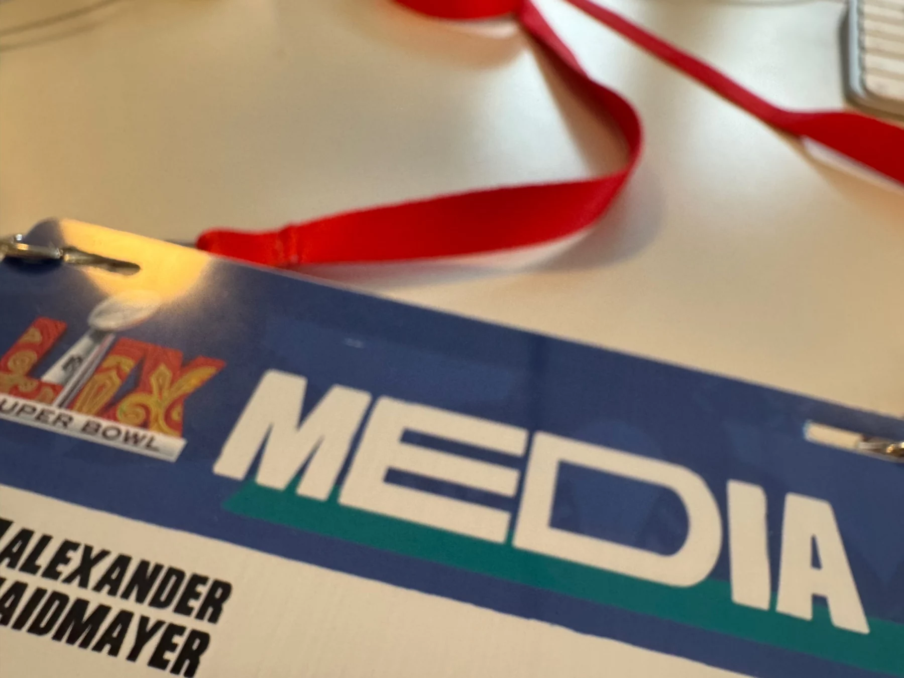 Super Bowl Media - Nahaufnahme eines Medienausweises des Super Bowl LIX auf einem Tisch. Der Ausweis zeigt links das ikonische Logo der Veranstaltung und in der Mitte deutlich das Wort „MEDIA“ und zieht damit die Aufmerksamkeit der Football-Fans auf sich. Ein rotes Schlüsselband schmiegt sich elegant an den Tisch, darunter ist teilweise „ALEXANDER MAIDMAYER“ zu sehen. Diese Beschreibung wurde mit der FootballR KI automatisch generiert.