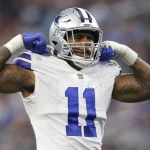 Micah Parsons, der die weiße Uniform der Dallas Cowboys mit der Nummer 11 trägt, lässt während eines Spiels selbstbewusst seine Muskeln spielen. Sein silberner Helm trägt einen blauen Streifen, und beide Arme sind mit Tattoos geschmückt. Hinter ihm bilden das verschwommene Publikum und die Stadionlichter eine elektrisierende Kulisse. Diese Beschreibung wurde mit der FootballR KI automatisch generiert.