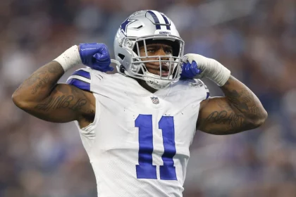 Micah Parsons, der die weiße Uniform der Dallas Cowboys mit der Nummer 11 trägt, lässt während eines Spiels selbstbewusst seine Muskeln spielen. Sein silberner Helm trägt einen blauen Streifen, und beide Arme sind mit Tattoos geschmückt. Hinter ihm bilden das verschwommene Publikum und die Stadionlichter eine elektrisierende Kulisse. Diese Beschreibung wurde mit der FootballR KI automatisch generiert.
