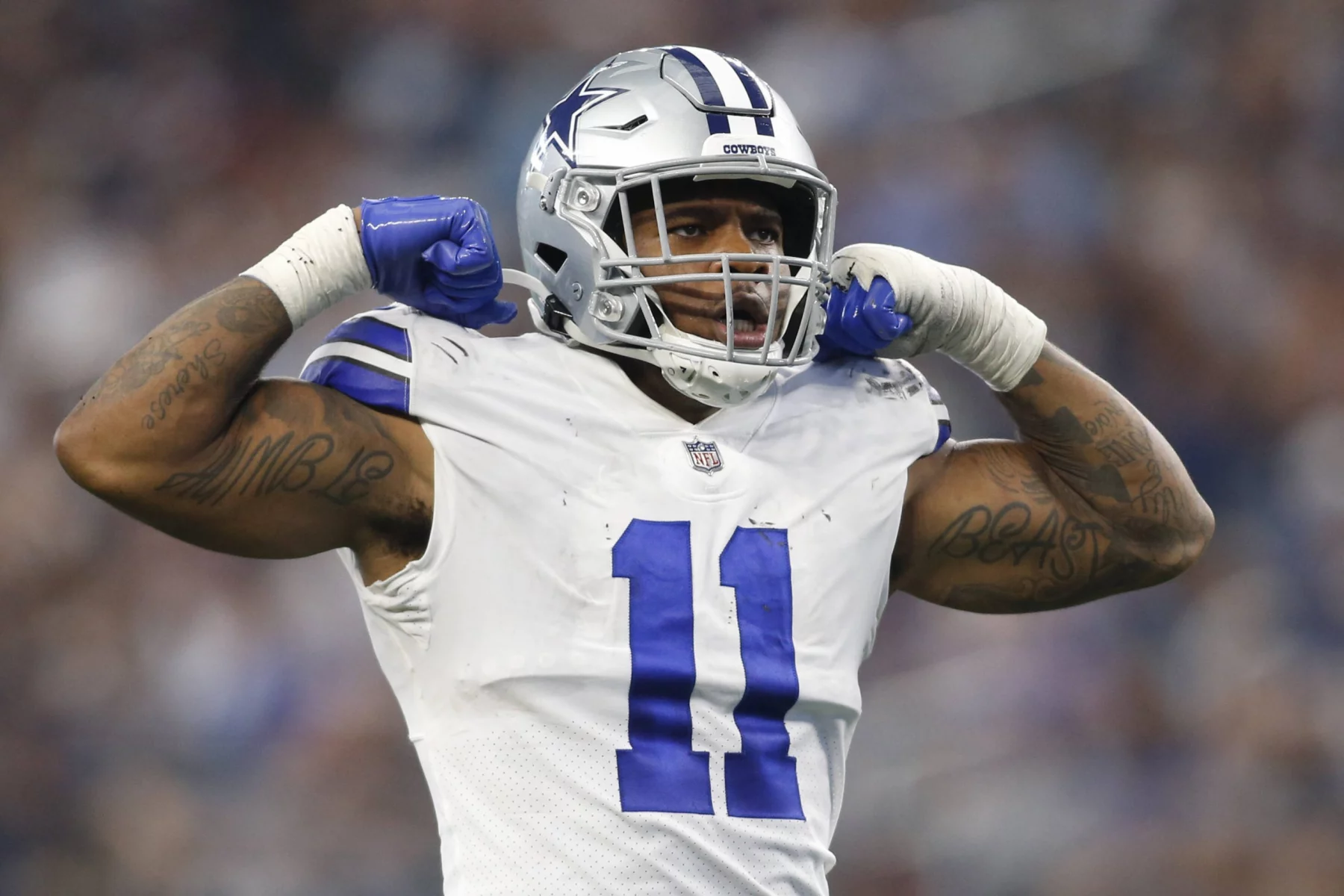 Micah Parsons, der die weiße Uniform der Dallas Cowboys mit der Nummer 11 trägt, lässt während eines Spiels selbstbewusst seine Muskeln spielen. Sein silberner Helm trägt einen blauen Streifen, und beide Arme sind mit Tattoos geschmückt. Hinter ihm bilden das verschwommene Publikum und die Stadionlichter eine elektrisierende Kulisse. Diese Beschreibung wurde mit der FootballR KI automatisch generiert.