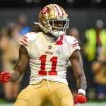 Ein Footballspieler, der Brandon Aiyuk ähnelt, trägt ein Trikot der San Francisco 49ers mit der Nummer 11 und steht auf dem Spielfeld. Seine langen Haare lugen unter einem goldenen Helm hervor, er hat die Fäuste geballt, und ein angespannter Gesichtsausdruck ziert sein Gesicht. Im Hintergrund sind ein Schiedsrichter und verschwommene Zuschauer zu sehen. Diese Beschreibung wurde mit der FootballR KI automatisch generiert.