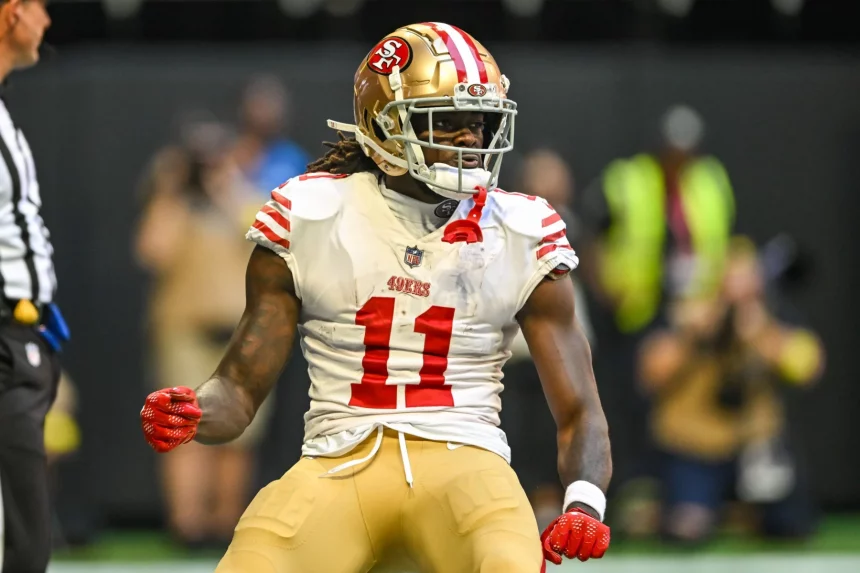 Ein Footballspieler, der Brandon Aiyuk ähnelt, trägt ein Trikot der San Francisco 49ers mit der Nummer 11 und steht auf dem Spielfeld. Seine langen Haare lugen unter einem goldenen Helm hervor, er hat die Fäuste geballt, und ein angespannter Gesichtsausdruck ziert sein Gesicht. Im Hintergrund sind ein Schiedsrichter und verschwommene Zuschauer zu sehen. Diese Beschreibung wurde mit der FootballR KI automatisch generiert.