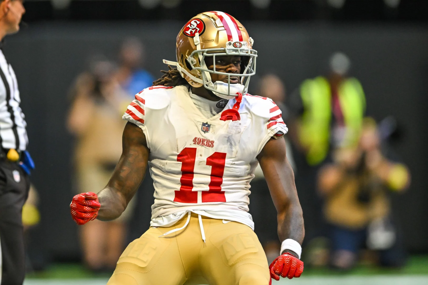 Ein Footballspieler, der Brandon Aiyuk ähnelt, trägt ein Trikot der San Francisco 49ers mit der Nummer 11 und steht auf dem Spielfeld. Seine langen Haare lugen unter einem goldenen Helm hervor, er hat die Fäuste geballt, und ein angespannter Gesichtsausdruck ziert sein Gesicht. Im Hintergrund sind ein Schiedsrichter und verschwommene Zuschauer zu sehen. Diese Beschreibung wurde mit der FootballR KI automatisch generiert.