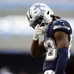 Ceedee Lamb im Trikot der Dallas Cowboys steht auf dem Spielfeld und vollführt die klassische "Nasenwischer" Geste. Er trägt ein marineblaues Trikot mit weißen Akzenten, die Nummer 88, einen silbernen Helm und weiße Handschuhe und rückt konzentriert seinen Helm zurecht – ein Zeichen seiner Hingabe an die NFL-Regeln. Diese Beschreibung wurde mit der FootballR KI automatisch generiert.